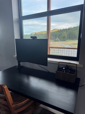A desk with a computer monitor, a basket, and a window with a view of a forest and a road.