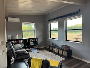 A living room with a black leather sofa, a coffee table, a bookshelf, and large windows offering a view of a field.