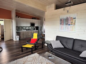 A modern living room with a black leather sofa, a glass coffee table, and a dining area with wooden cabinets and a tiled backsplash.
