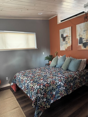 A bedroom with a floral bedspread, a window, a plant, and abstract wall art.