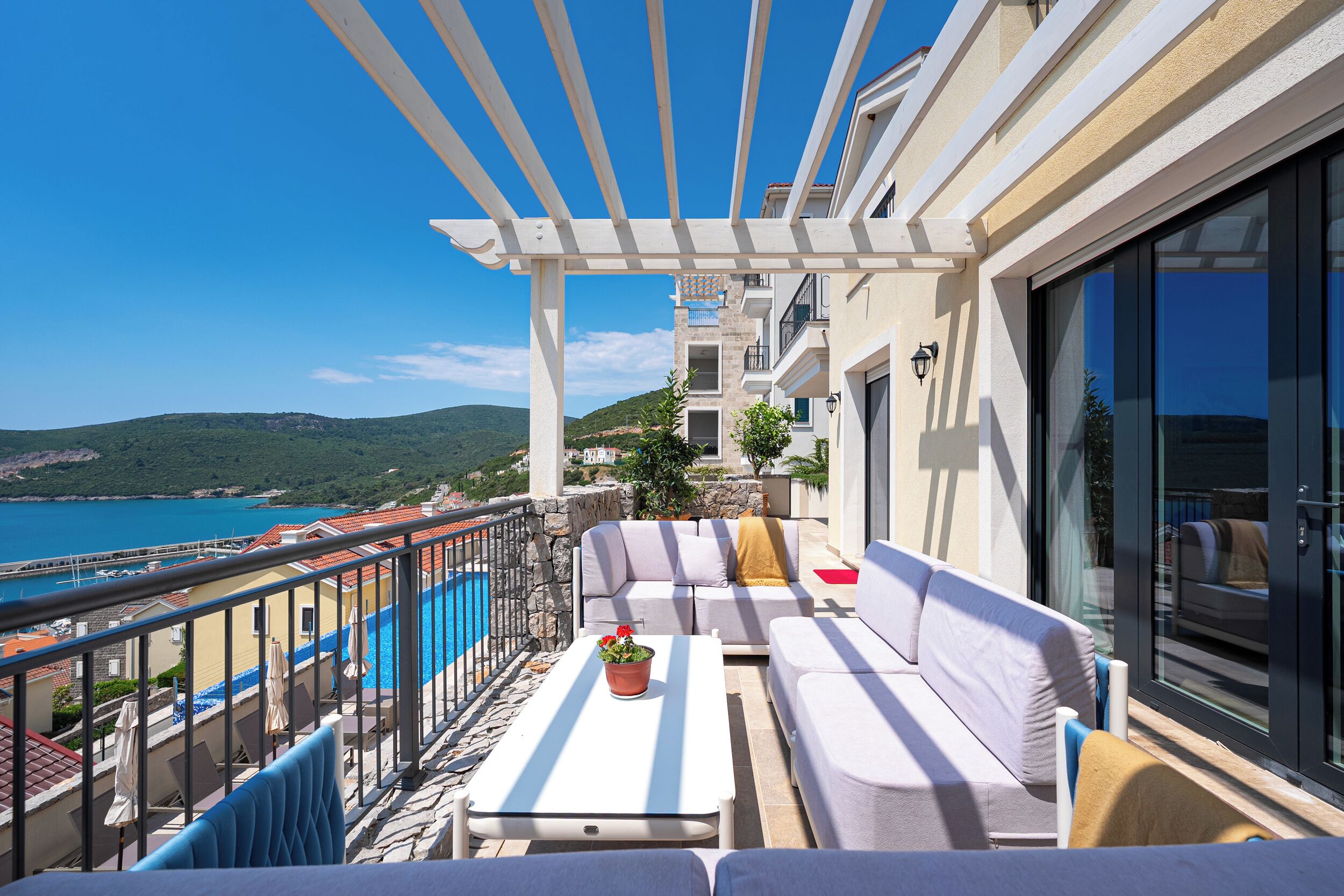 Un balcon avec un canapé blanc, une petite table sur laquelle est posée une plante en pot, et une vue sur la mer et les collines.