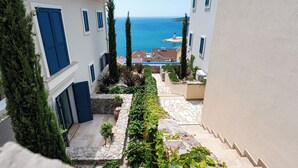 Eine enge Gasse mit Steinstufen, die zu einem Gebäude mit blauen Fensterläden führen, mit Blick auf das Meer und einem Leuchtturm in der Ferne.