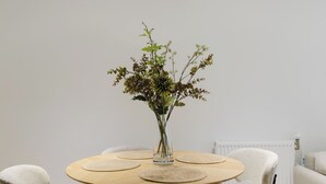 Una mesa redonda de madera con un florero de vidrio transparente que contiene follaje verde, rodeada por cuatro individuales beige.