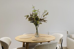 A round wooden table with a clear glass vase containing green foliage, surrounded by four beige placemats. - Stylish 1BDR Flat with Private Balcony in Vauxhall (London)