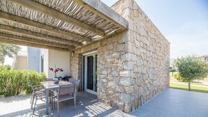 En mur af natursten, en overdækket terrasse med et bord og stole, og en glasdør ind til bygningen.