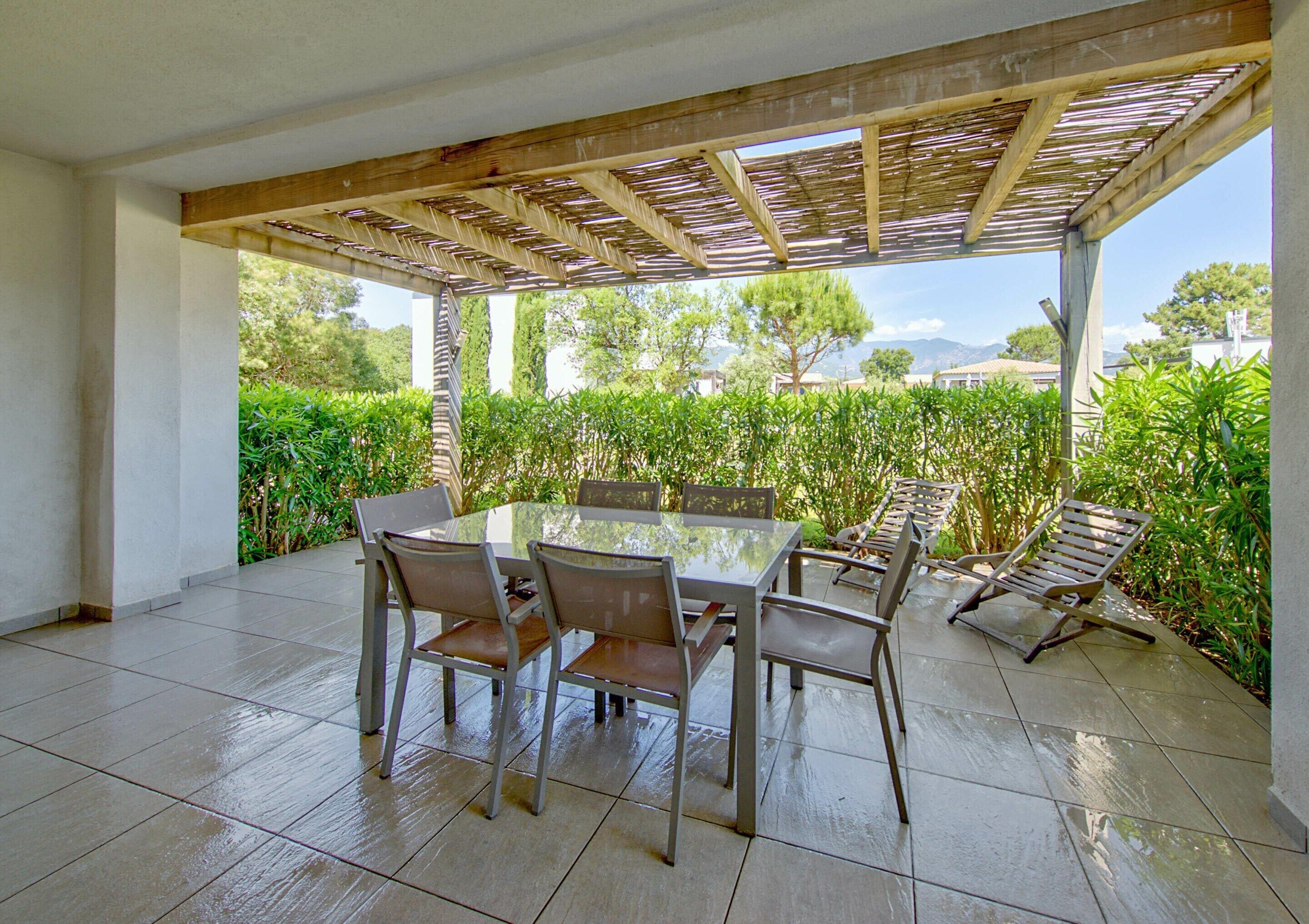 Eine Terrasse im Freien mit einem Tisch und Stühlen mit Glasplatte, umgeben von Grünpflanzen und einer Holzpergola.