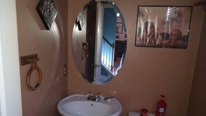 Une salle de bain avec un miroir rond, un lavabo, des toilettes, un extincteur et un tableau encadré au mur.