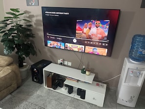 A living room with a flat-screen TV mounted on the wall, a potted plant, a beige sofa, a white entertainment unit with electronic devices, and a water dispenser.