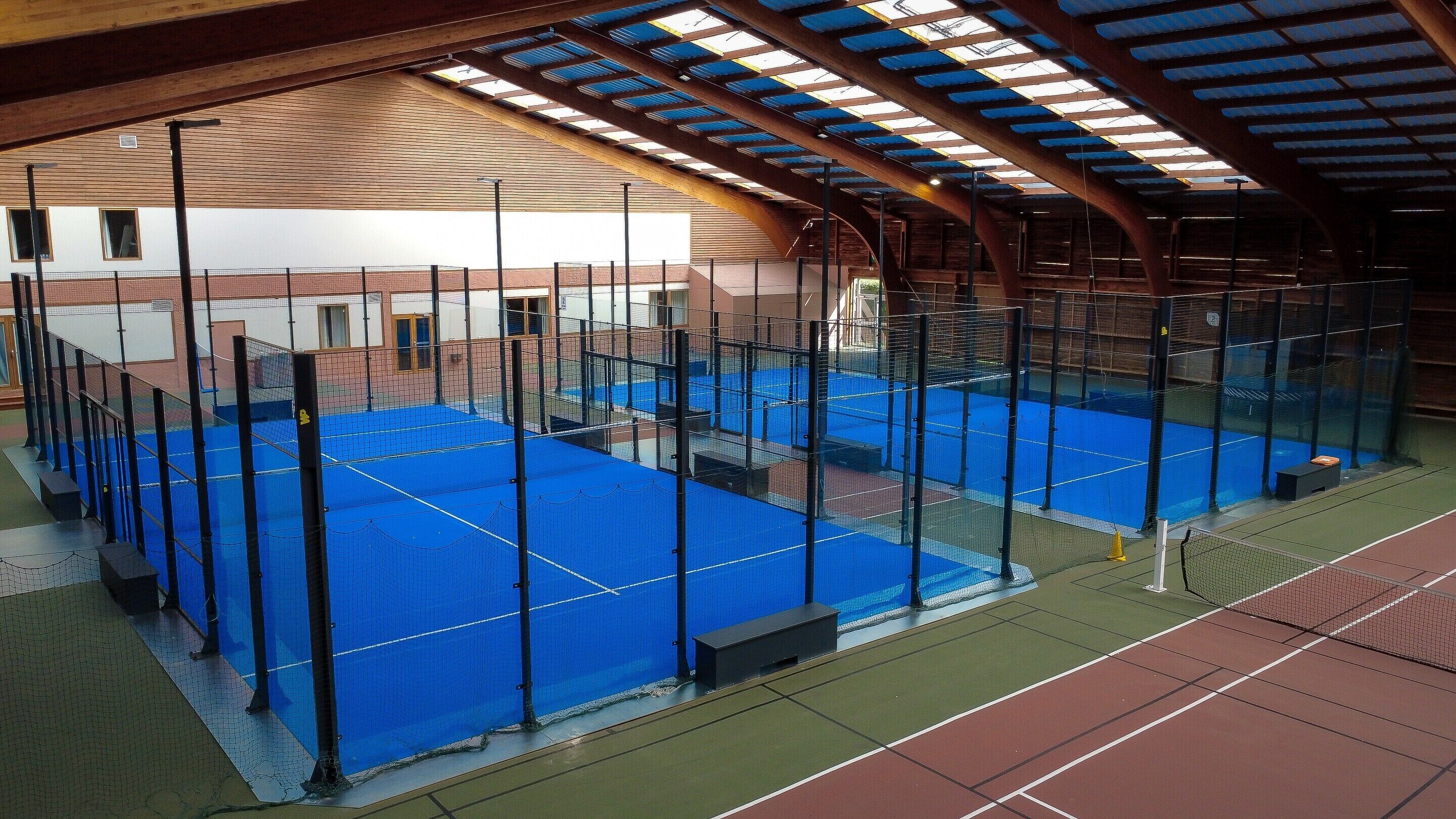 Impianto sportivo interno con diverse piste da tennis, un tetto coperto e un edificio in mattoni sullo sfondo.