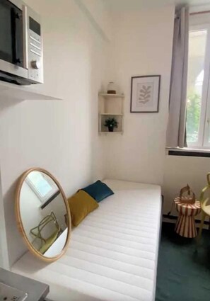 Ein kleines Zimmer mit einem Einzelbett, einem Nachttisch, einem Spiegel, einem Fenster mit Vorhängen, einem Regal mit einer Pflanze und einem Bilderrahmen an der Wand.