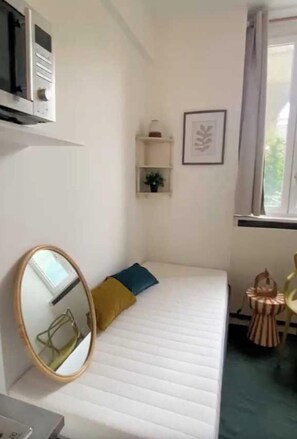 Ein kleines Zimmer mit einem Einzelbett, einem Nachttisch, einem Spiegel, einem Fenster mit Vorhängen, einem Regal mit einer Pflanze und einem Bilderrahmen an der Wand.