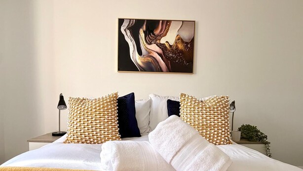 A neatly made bed with white linens, two decorative pillows, a gold throw blanket, and a wall-mounted abstract painting. - Swan View House in Littleport (Littleport)