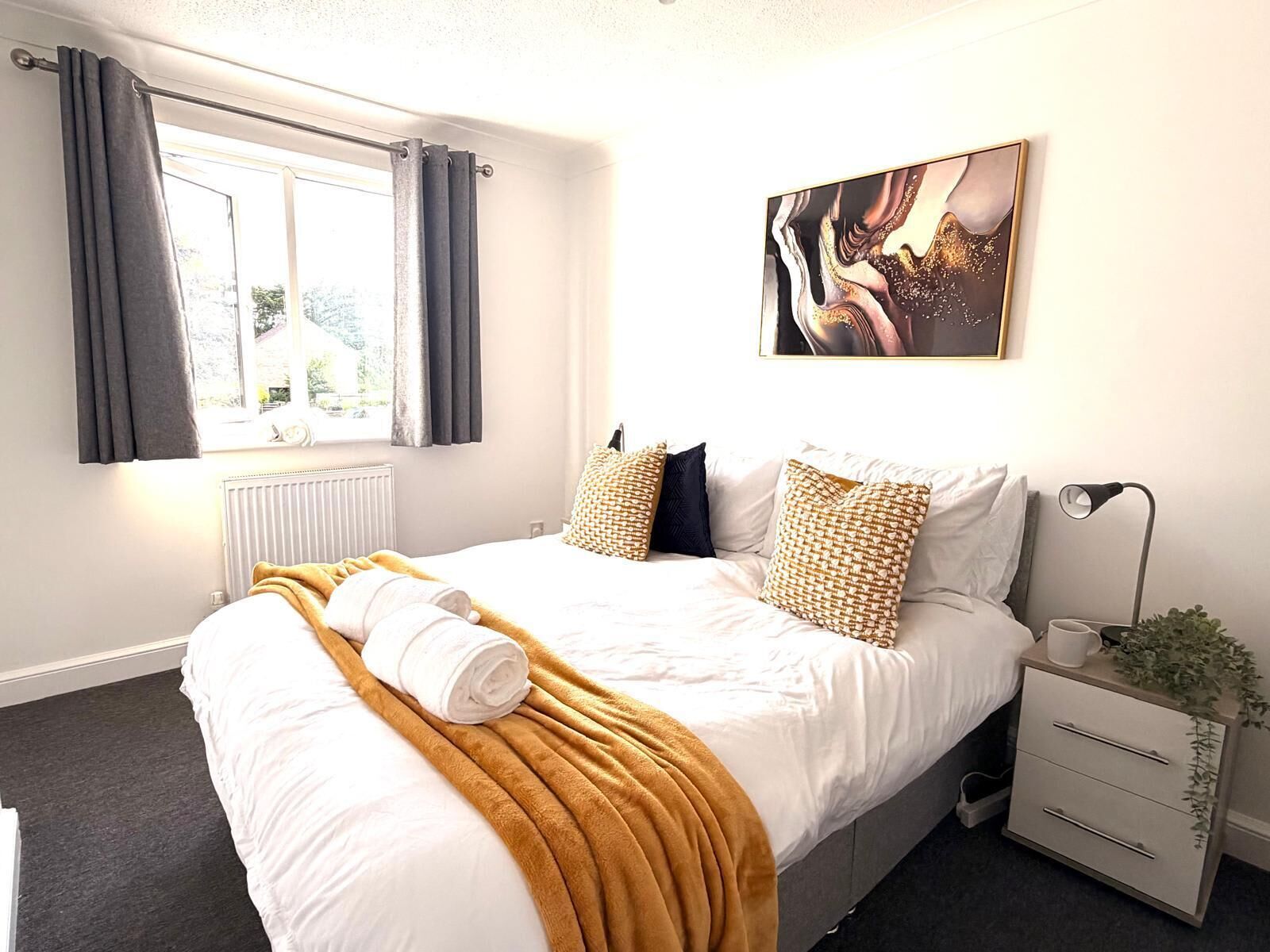 A hotel room with a bed, a window with curtains, a radiator, a bedside table with a lamp, and a wall-mounted artwork.. Swan View House in Littleport