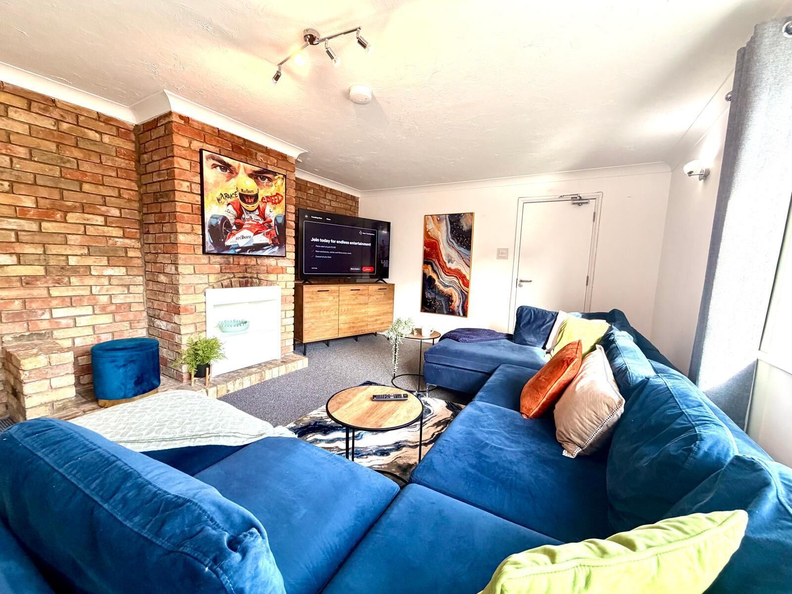 Un salon moderne avec un mur d’accent en briques, un ensemble de canapés bleus, un téléviseur à écran plat et une cheminée.