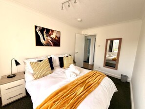A bedroom with a bed, bedside table, lamp, artwork, mirror, and a door leading to another room. - Swan View House in Littleport (Littleport)