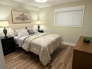 Ein Schlafzimmer mit einem Bett, Nachttischen, einer Lampe, einem Fenster mit Jalousien und einem Bild an der Wand.