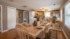 A dining area with a table set for six, wooden chairs, a ceiling fan, and a kitchen in the background. - Sunrise Retreat Private Pool & Hot Tub. Golfers Delight. Steps to the beach. (North Myrtle Beach)