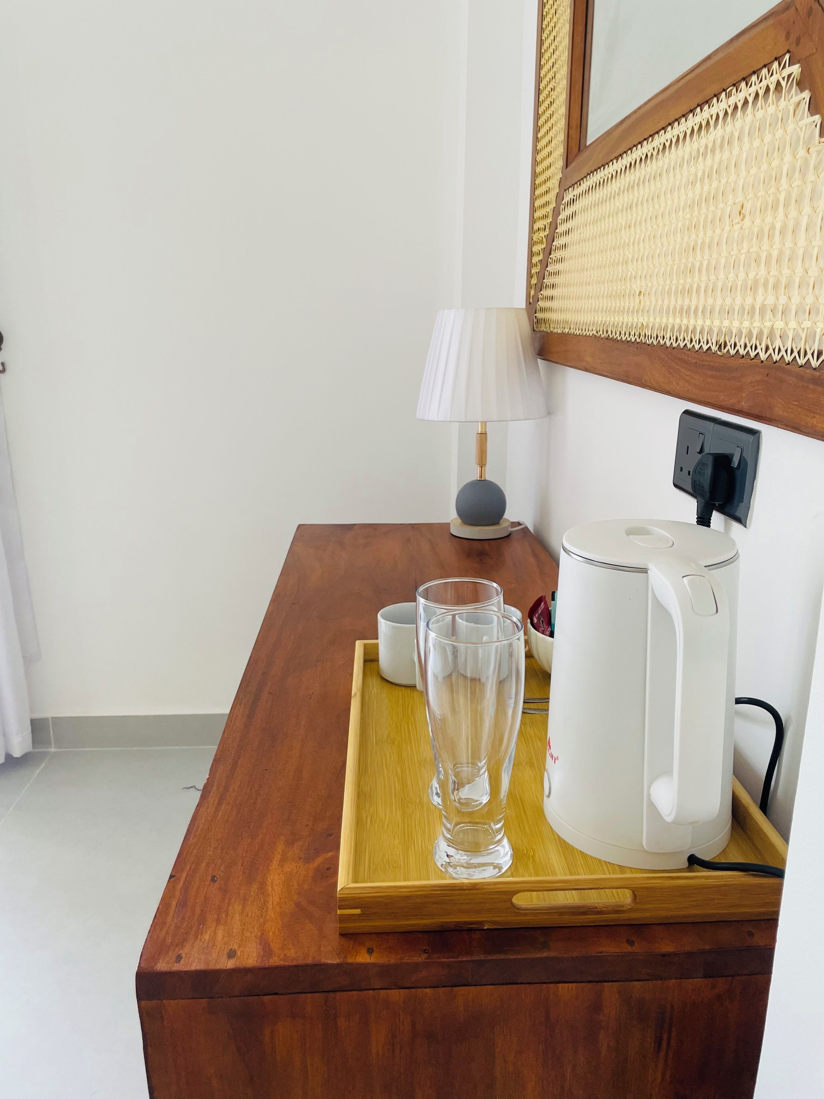 Une table de chevet en bois sur laquelle se trouvent une bouilloire blanche, un verre transparent et une tasse blanche, le tout posé sur un plateau.