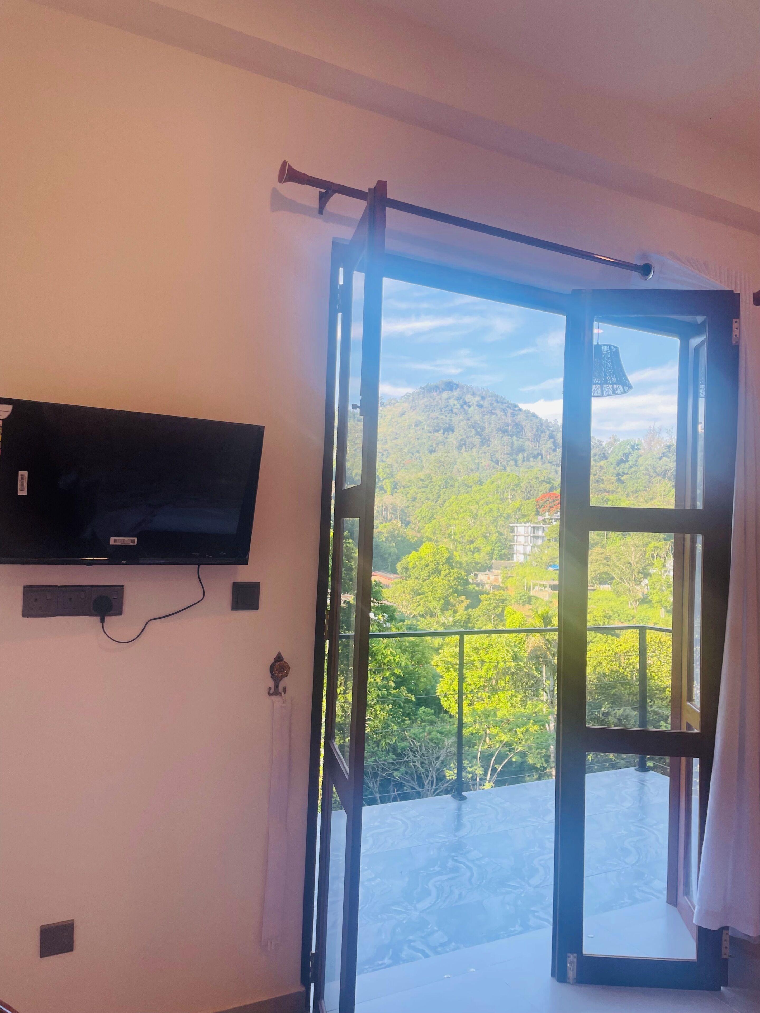 A room with a television mounted on the wall, a sliding glass door leading to an outdoor balcony with a view of a green landscape and hills.