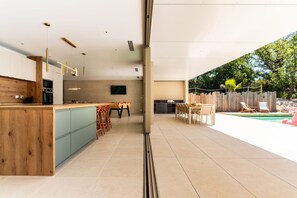 A modern kitchen with a wooden island, a dining area with chairs, and a poolside with lounge chairs. - Villa Joy Pool Sauna River Escape (Murviel-les-Beziers)