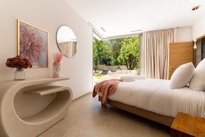 A modern bedroom with a large bed, a curved console table, a round mirror, and a view of an outdoor area with greenery. - Villa Joy Pool Sauna River Escape (Murviel-les-Beziers)
