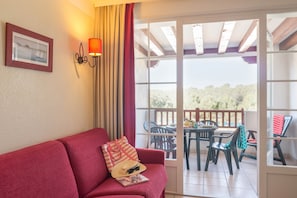Un canapé rouge avec un magazine et un chapeau, une table avec des chaises, et une vue sur les arbres à travers une porte vitrée.