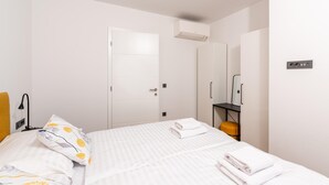 Un dormitorio con cama, paredes blancas, una puerta, un equipo de aire acondicionado, un espejo y una pequeña mesa con silla.