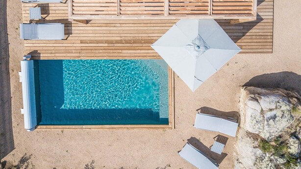 Vista aérea de una zona de piscina con terraza de madera, sillones de descanso y sombrillas.