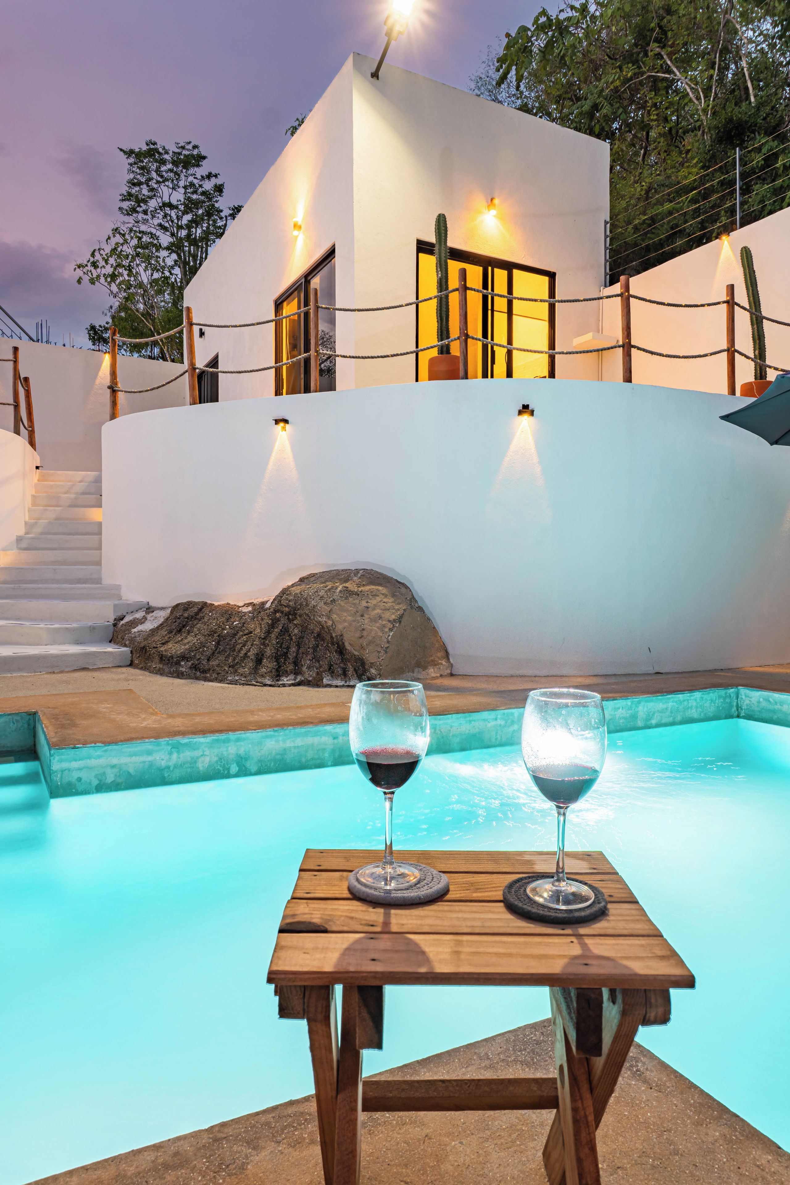 Ein Tisch am Pool mit zwei Gläsern Rotwein, ein modernes weißes Gebäude mit einer Treppe und eine Kakteenpflanze.