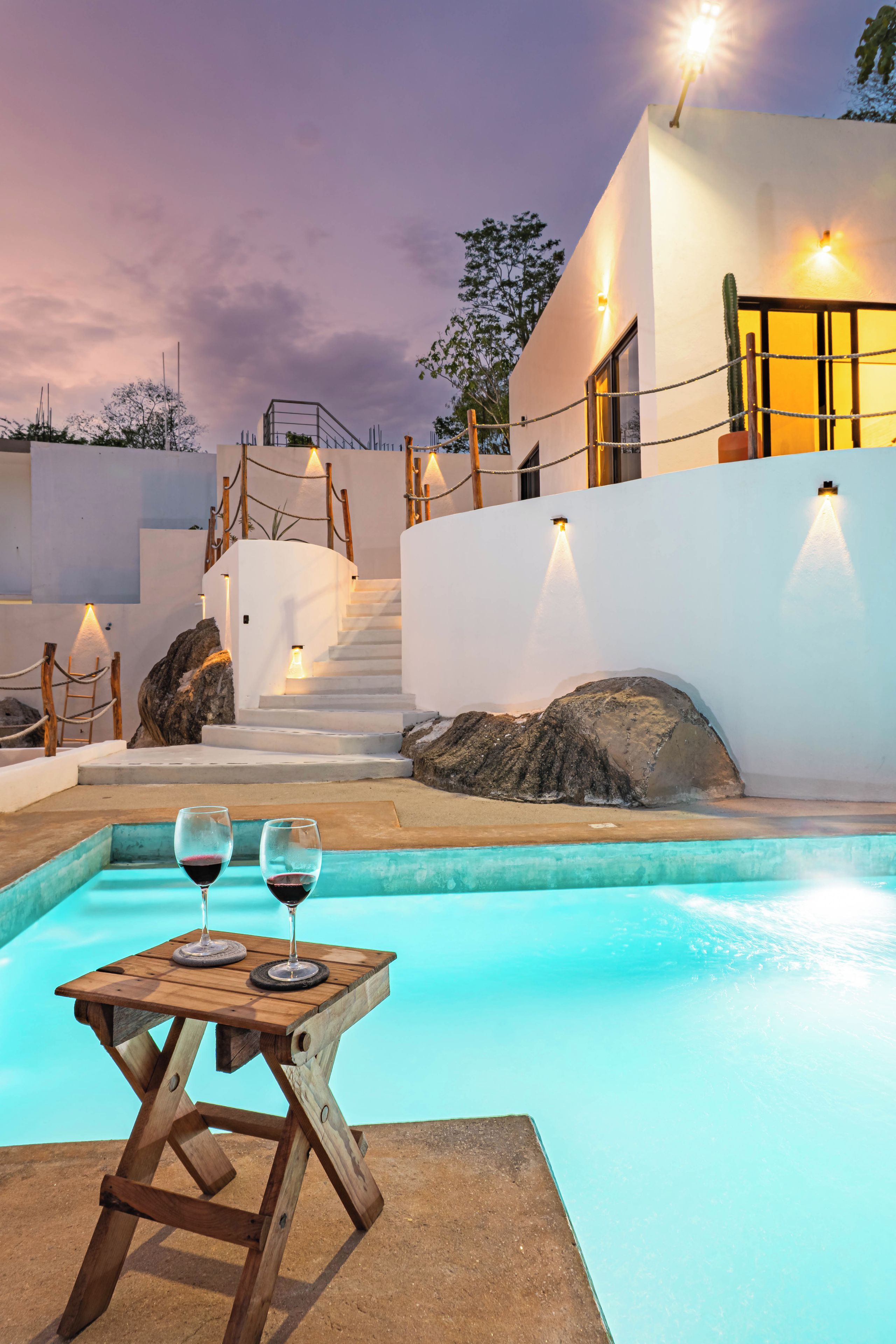 Zona de piscina moderna al aire libre con una mesa de madera y dos copas de vino tinto, rodeada de paredes blancas y una escalera que conduce a un edificio.