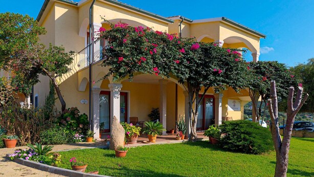 A yellow building with a covered entrance, potted plants, and a well-maintained lawn. - Spacious Beach Apartment for 6 (Badesi)