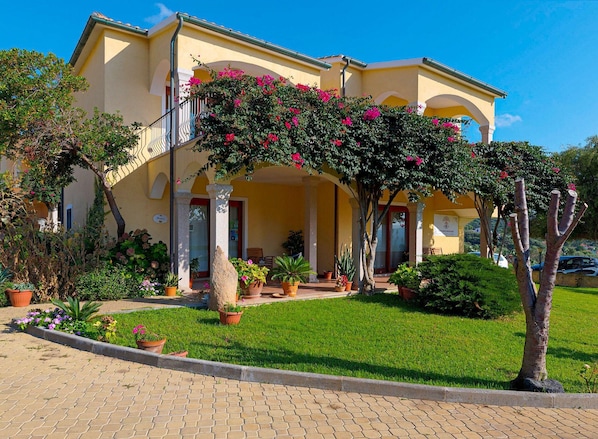 A yellow building with a covered entrance, potted plants, and a well-maintained lawn. - Spacious Beach Apartment for 6 (Badesi)