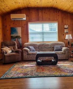 A cozy living room with a wooden ceiling, a sofa, a chair, a coffee table, and a large window with blinds.