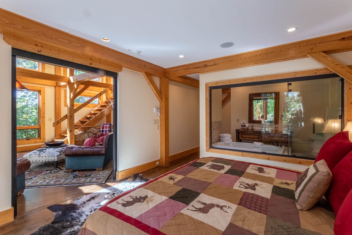 A spacious room with a large window, a wooden staircase, and a cozy seating area with a quilted rug.