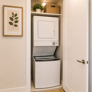 Une petite buanderie blanche, dotée d’un lave-linge et d’un sèche-linge encastrés, d’une estampe botanique encadrée et d’un panier en osier posé sur une étagère.