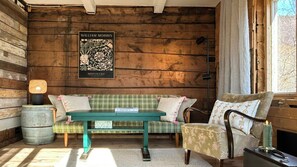 A cozy room with a wooden wall, a framed poster, a green sofa, a patterned armchair, a small table with a lamp, and a window with curtains.