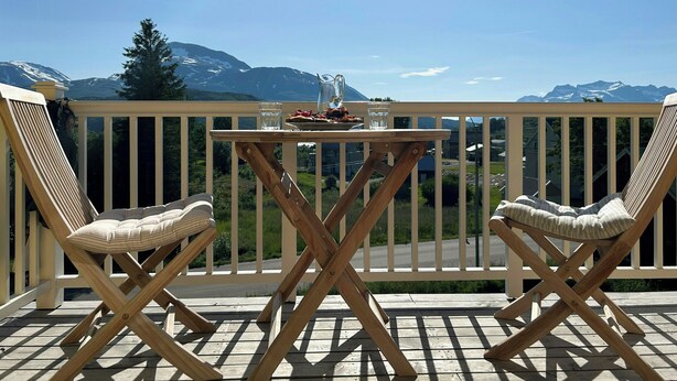 Een houten terras met twee comfortabele stoelen en een tafel, met uitzicht op een bergachtig landschap.