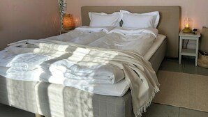 Een net opgemaakt bed met een witte deken en een beige plaid, een bedlampje en een klein tafeltje met een vaas.