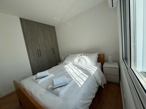 Ein ordentlich bezogenes Bett mit Handtüchern, ein Holzschrank, ein Nachttisch mit Lampe und ein Fenster mit Vorhängen.