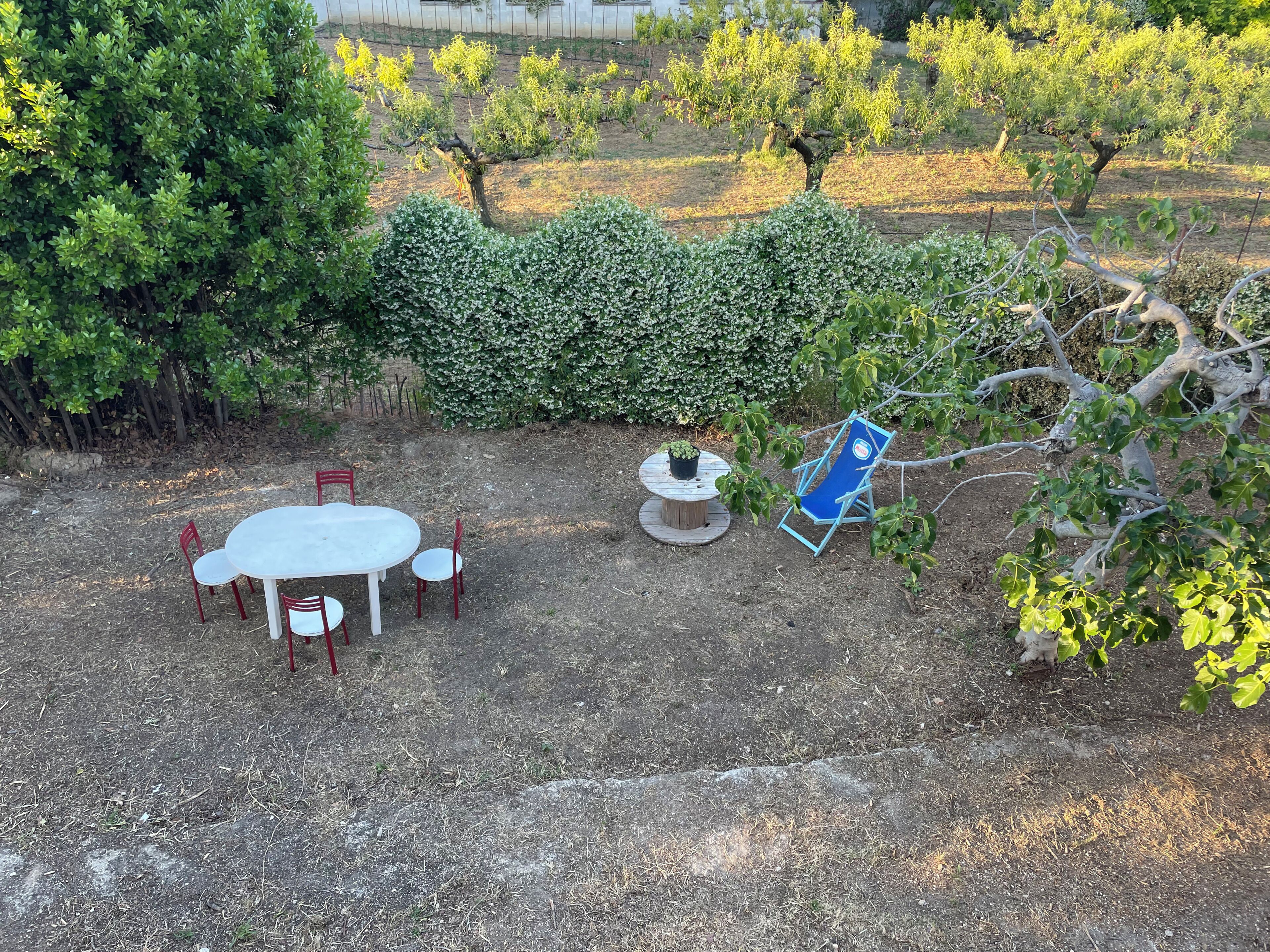 Eine Terrasse mit rundem Tisch und Stühlen, ein blauer Klappstuhl und ein Hecken.