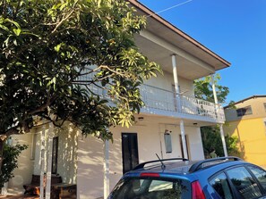 Um edifício de dois andares com uma varanda, um carro estacionado e uma árvore em primeiro plano.