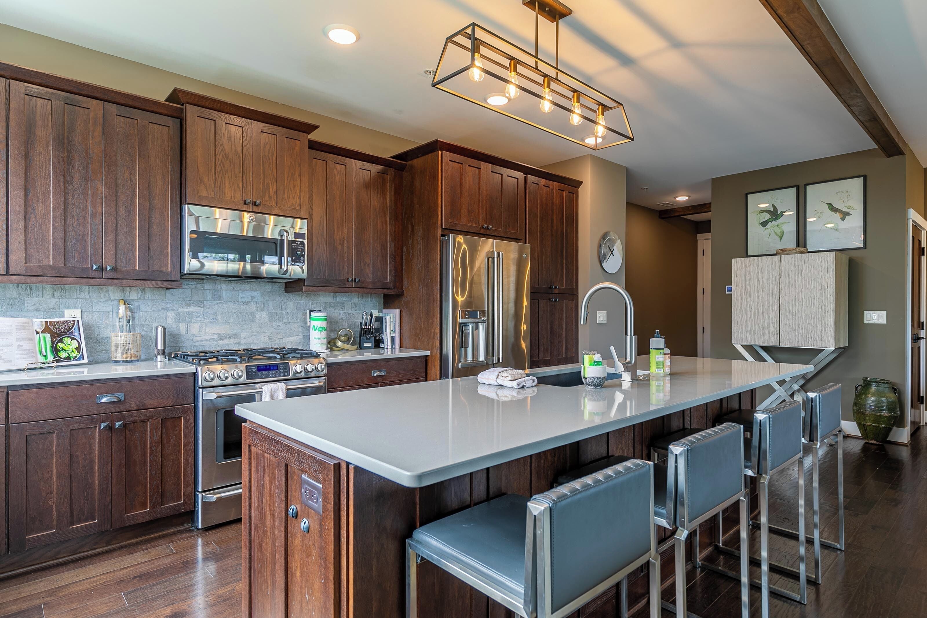 A modern kitchen with wooden cabinets, stainless steel appliances, a central island with bar stools, and a decorative light fixture.
