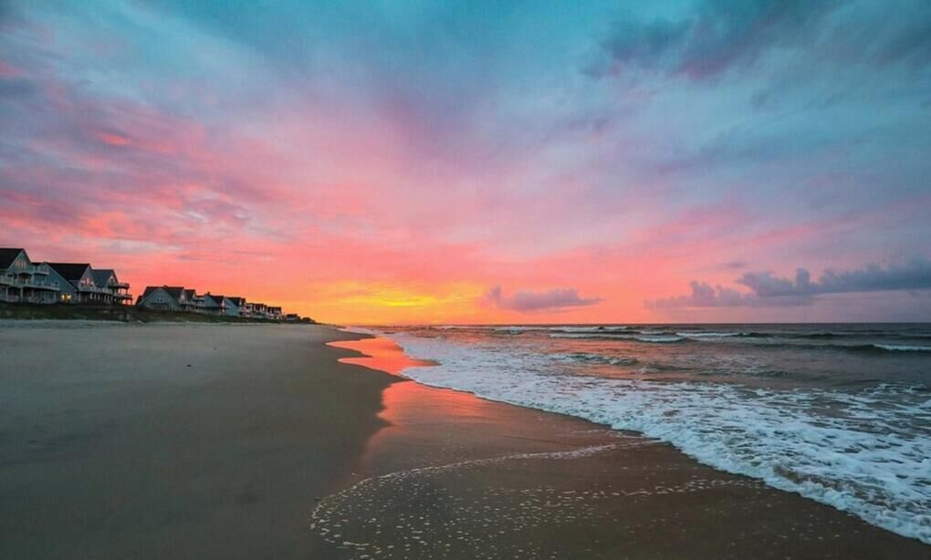 A beach with houses along the shore, a sunset, and waves.