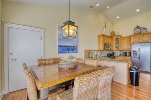 A dining area with a wooden table and wicker chairs, a kitchen with wooden cabinets and stainless steel appliances, and a painting of a seascape on the wall.