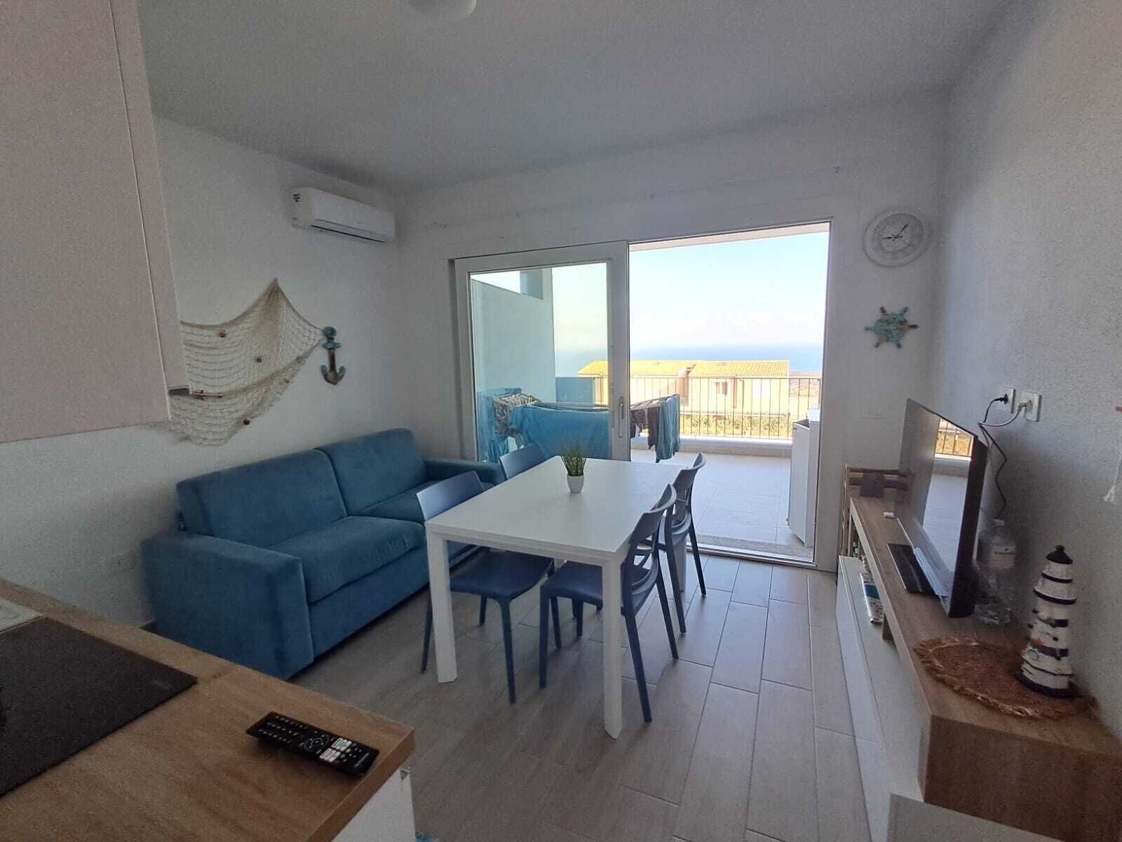 Habitación con sofá azul, una mesa blanca y sillas, televisor y balcón con vistas.