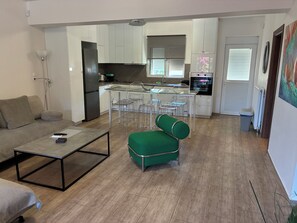 Una sala de estar moderna con un sofá, una mesa de centro y un sillón verde. Se ve una cocina con armarios blancos y una zona de comedor con sillas transparentes al fondo.