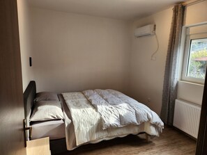 A single bed with a headboard, a window with curtains, an air conditioning unit, and a radiator.