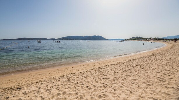 Ein Strand mit klarem Wasser, Booten und Bergen im Hintergrund.