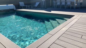 Ein rechteckiger Swimmingpool mit klarem Wasser, umgeben von einer Holzterrasse und weißen Liegestühlen.