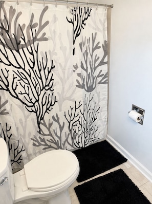 A bathroom with a toilet, a shower curtain with a coral pattern, a black rug, and a toilet paper holder on the wall.